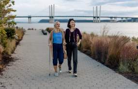 Two people are walking along the Riverwalk in Tarrytown. The Gov. Mario Cuomo Bridge is in the background.