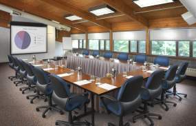 A meeting room with the tables arranged in a U-shape. The chairs are facing a screen with a pie graph on it.