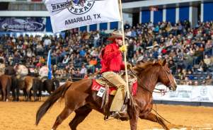 A cowboy wearing a red shirt, tan chaps, and a cowboy hat rides a galloping chestnut horse inside an indoor rodeo arena. He holds a large white flag that reads "West Texas Ranch Rodeo, Amarillo, TX," with a logo in the center. The background shows a packed audience in the stands and several other riders on horseback in the arena. The dirt-covered floor and western attire emphasize the rodeo setting.