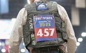 A rodeo competitor leans against a blue fence inside an arena, wearing a black cowboy hat, leather vest, and numbered back tag for the Tri-State Fair & Rodeo in Amarillo, Texas, held September 13–21, 2024. The lights blur in the background as the rider looks on, chaps swaying at his sides, capturing the anticipation and grit of rodeo life in the Panhandle.