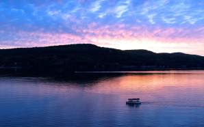 A lake with hills in the background with a boat and a blue, pink, and orange sunset in the sky and reflecting in the water.