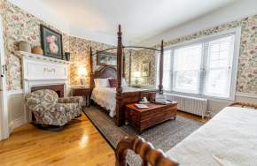 Interior of a bedroom with floral wallpaper in an historic inn with a full bed with a wooden headboard, a sitting chair, and a fireplace.