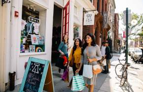 Three women with shopping bags and purses walking down a main street sidewalk