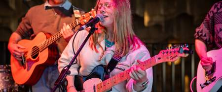 A young woman singing and playing the guitar in a lit up venue.