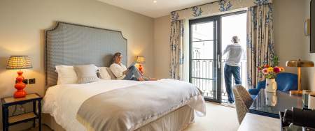 A man and a woman in a hotel room. Woman reading on the bed, man looking out of the window.