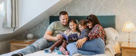A man, woman and two girls reading a story book on a bed.