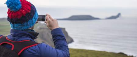 A man wearing a bobble hat taking a photo of Worm's Head.