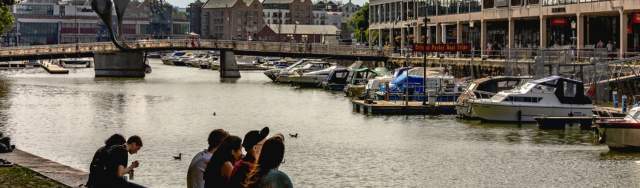 People sitting on the Bristol Harbourside with Pero's Bridge and the cranes outside M Shed in the background