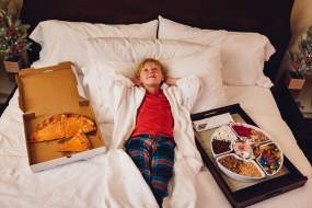 A boy lays on a hotel bed at the Westin Poinsett in Greenville, SC wearing a white fluffy robe with cheese pizza on one side of him and an ice cream sundae and lots of toppings on the other.