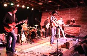 Four musicians play guitar and one plays drums on a stage