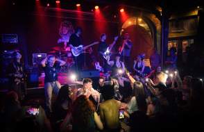Concert goers hold up phone flashlights as a band plays on stage