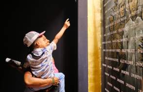 African American man holding a young child in front of the plaque with engraved names of the Clotilda, the Last American Slave Ship