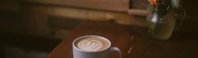 A latte served in a white cup sits on a table next to a vase with orange and yellow flowers.