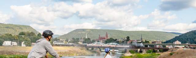 Adult and child riding bicycles along the C&O Canal Towpath in Cumberland, Maryland, with a river, bridge, downtown buildings, and green mountains and Western Maryland Scenic Railroad steam engine in the background under a blue sky.