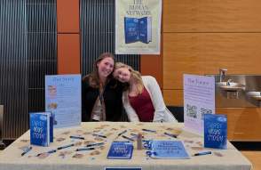 Two woman sitting at table with books and sign saying The Human Network