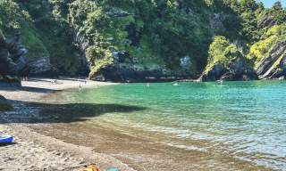 A serene cove with turquoise waters, surrounded by lush green cliffs. People are kayaking and relaxing on the sandy shore under a clear blue sky.