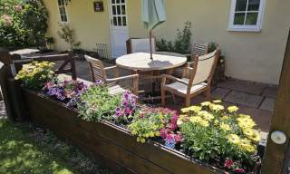 A cosy patio with wooden chairs and a round table shaded by a green umbrella. Vibrant yellow and pink flowers in a raised bed, backed by a yellow cottage.