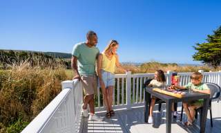 A family enjoys a sunny day on a deck by the beach. Two children sit at a table with drinks and snacks. A couple stands nearby, smiling and talking. Lush greenery and a clear blue sky create a relaxed, joyful ambience.