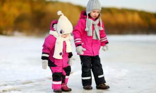 Two children in pink snowsuits, hats, and scarves walk on a snowy path. The scene is bright and playful, with soft trees blurred in the background.