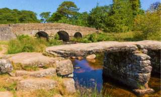 A picturesque stone bridge with three arches spans a gentle stream, surrounded by lush green trees and rocks under a clear blue sky. Calm and serene atmosphere.