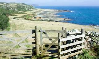 A wooden gate opens to a coastal path with distant green hills and a vast, calm blue sea. The scene conveys a serene, inviting atmosphere.§