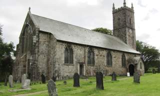 A historic stone church with tall arched windows and a square bell tower stands amidst a green graveyard. The scene feels tranquil under a cloudy sky.