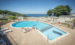 Outdoor pool with loungers, people sunbathing, and a colourful inflatable under a clear blue sky. Mobile homes and sea view in the background.