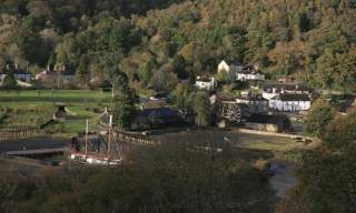 A scenic village nestled in a lush, forested valley. Features quaint cottages, a waterwheel, open green fields, and a docked ship, evoking tranquillity.