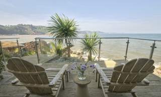 Two wooden lounge chairs on a deck overlook a serene sea view, framed by tropical plants and a glass railing, with a vase of purple flowers on a small table between them.