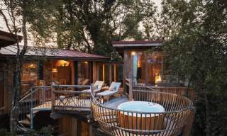 Treehouse retreat at sunset with large windows, cosy interior lighting, chaise lounges, and a hot tub on a wooden deck surrounded by lush trees.