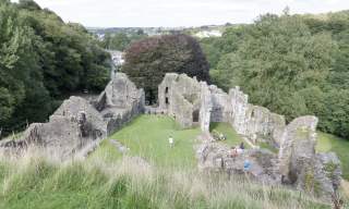 Ancient stone ruins surrounded by lush green trees and grass, with people exploring the site. The scene conveys a sense of history and tranquillity.