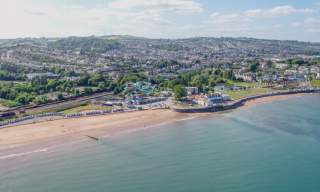 Aerial view of a coastal town with a sandy beach lined with blue beach huts. The sea is calm, and a scenic hillside town is in the background. The tone is peaceful.