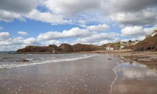 Coastal scene with rocky cliffs, sandy beach, and gentle waves under a sky with scattered clouds. Small houses and a boat are visible in the distance. Peaceful.