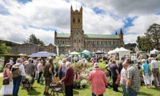 Crowded outdoor garden festival in front of a large, historic cathedral. People browse plant stalls under partly cloudy skies, conveying a lively atmosphere.