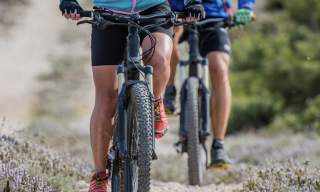 Two people riding mountain bikes on a rocky trail. They wear colourful jackets, shorts, and gloves. The background is blurred with greenery, conveying motion and adventure.