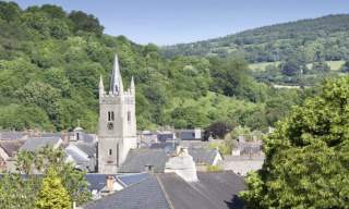 A scenic view of a quaint village, dominated by a stone church tower with a spire, surrounded by lush green hills under a clear blue sky.
