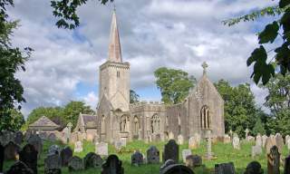 Historic stone church with a tall spire set amid a graveyard of weathered tombstones. Lush greenery surrounds, under a partly cloudy sky. Serene atmosphere.