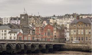 Historic townscape featuring a stone bridge, Victorian buildings, and a clock tower. The overcast sky and muted colors create a nostalgic atmosphere.
