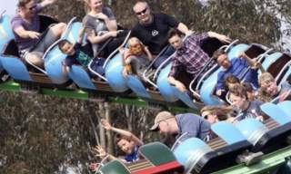 People are riding a roller coaster, showing excitement and thrill. The blue cars tilt as they speed down the track against a backdrop of trees.