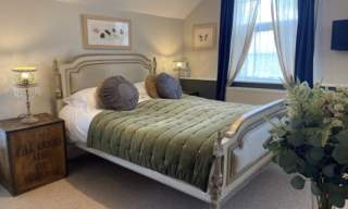 Cosy bedroom with vintage bed, green quilt, and round pillows. Soft lamp lighting, floral artwork, rustic crate, and fresh flowers create a calm ambience.