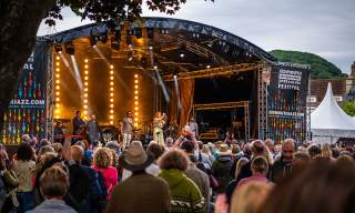 Outdoor music festival scene with a lively crowd watching performers on a lit stage. Banners read "Sidmouth Jazz and Blues Festival." Trees frame the view.