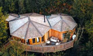 Aerial view of a wooden treehouse with hexagonal roofs, nestled among lush greenery. A cosy deck with chairs suggests relaxation and tranquility.