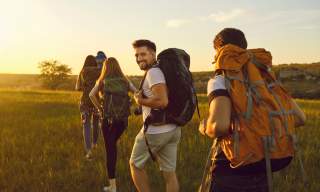 Four hikers with backpacks walk through a field at sunset. One glances back smiling, conveying a sense of adventure.