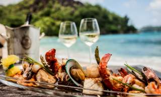 A plate of assorted seafood, including shrimp and mussels, on a table with two glasses of white wine. A scenic beach and lush greenery in the background.