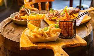 A rustic table holds a wooden board with crispy fries in a metal basket, calamari, and ribs with garnish. The scene is warm, cosy, and inviting.