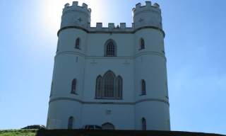 A white, castle-like tower with crenellations stands against a bright blue sky, the sun creating a halo effect. A car is parked on the grassy hill below.
