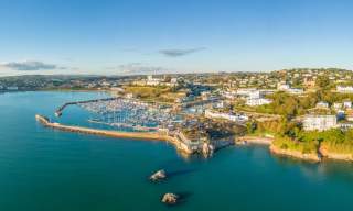 Aerial view of a coastal town with a marina full of boats, surrounded by lush greenery and buildings. The calm blue sea and clear sky create a serene atmosphere.