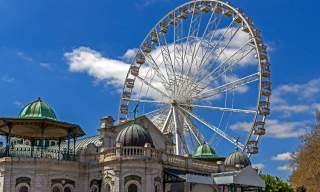 A large Ferris wheel rises behind an ornate historic building with domed green roofs under a bright blue sky with scattered clouds. A lively, whimsical atmosphere is conveyed.