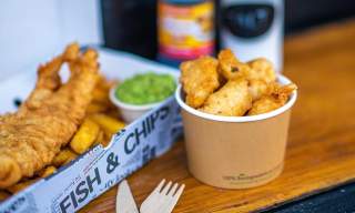 A hearty serving of fish and chips with mushy peas in a newspaper-lined box on a wooden table. A paper cup with fried snacks is nearby, evoking a warm, casual dining experience.