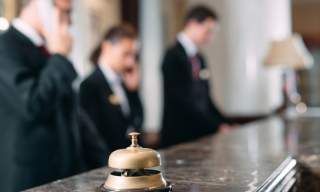 Hotel lobby with a focus on a service bell on a marble counter. In the background, blurred employees in formal attire use phones, conveying a busy atmosphere.